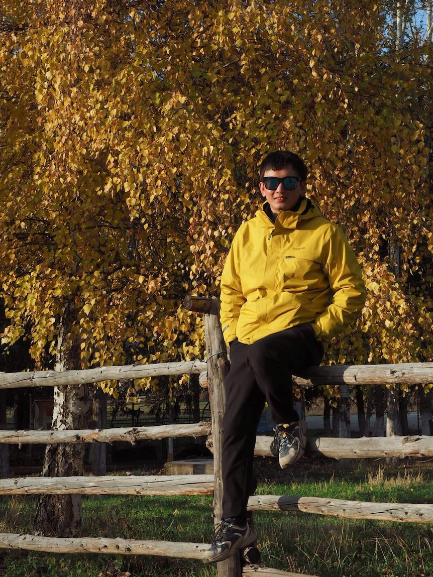 Shengwei Ye sits on a wooden fence with a large tree of yellow leaves behind him.