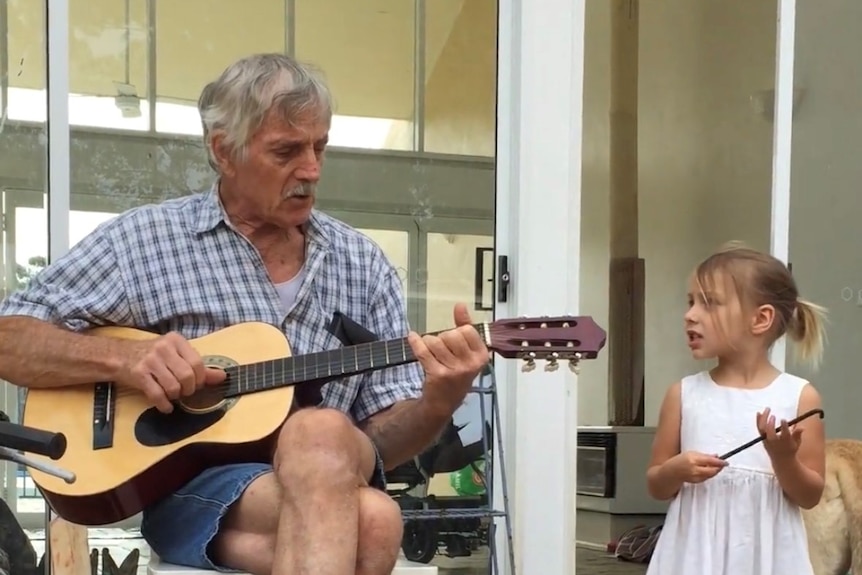 An older man sits playing a guitar to a young girl.
