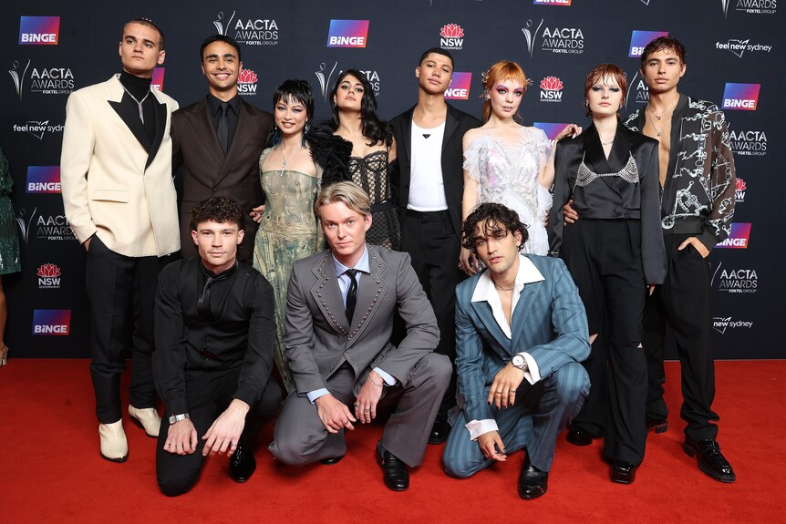 A group of 11 young people dressed in formal evening wear are assembled on a red carpet.