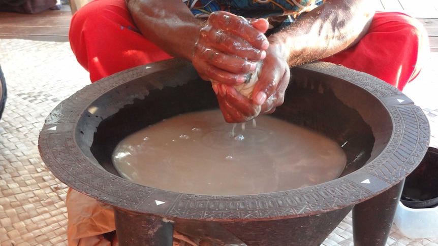 A man squats behind a large carved bowl with feet while making the traditional brown brew with his hands 