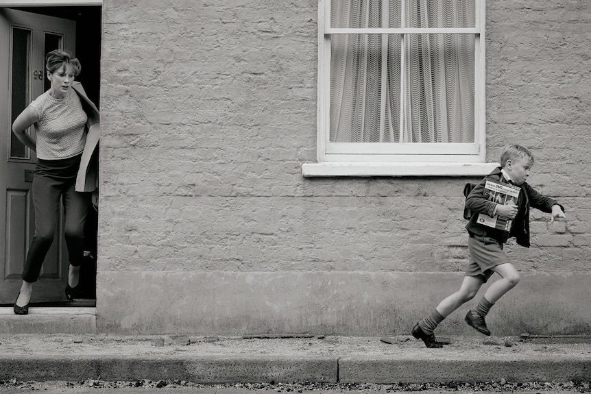 A black and white image of a middle-aged woman at her front door, chasing after a young boy running up the street