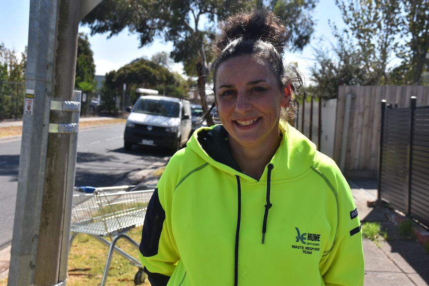 Candice Bowen wears a bright yellow hoodie with a Hume council logo and stands near a dumped trolley on a grassy footpath.