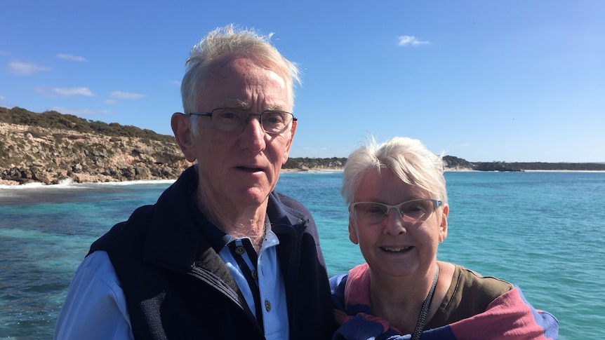 Ian and Claire White smile at the camera. They are outside and in front of a beautiful, blue ocean.