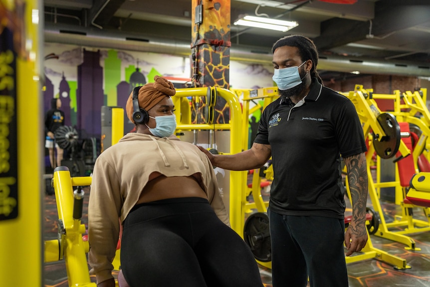 A young woman wearing head wrap, over-ear headphones and face mask works out indoors. A man also donning a face mask guides her