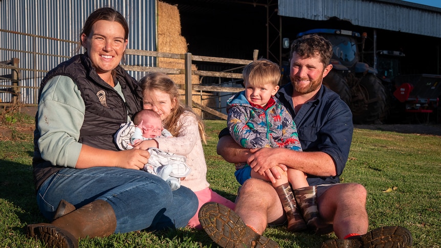 family of five including mother, father two toddlers and a baby sitting on grass in front of farm shed