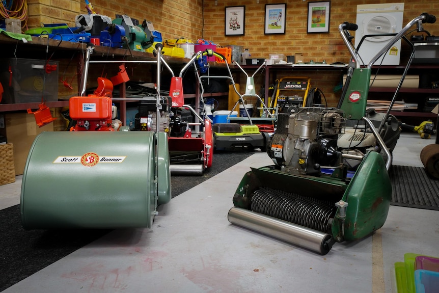 Six lawn mowers in a garage, at the front an old style cylinder mower and a lawn groomer