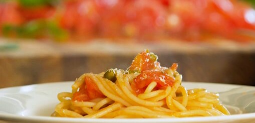 A spaghetti pasta with tomatoes and capers sits on a white plate, with chopped tomatoes on a wooden background in background blu