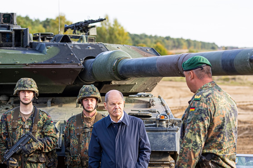 German leader stands beside three soldiers in front of Leopard 2 tank.
