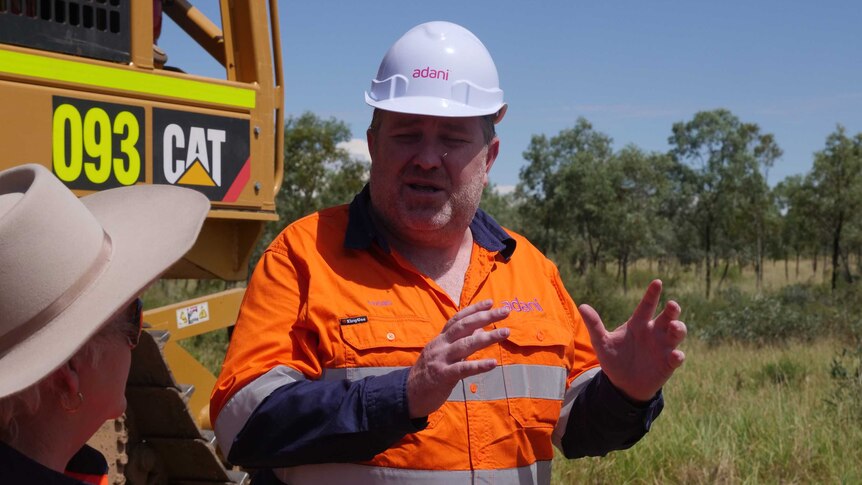 Adani Mining CEO Lucas Dow in fluorescent orange shirt and white hard hat speaking in front of trees and an orange tractor