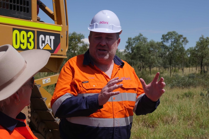 Adani Mining CEO Lucas Dow in fluorescent orange shirt and white hard hat speaking in front of trees and an orange tractor