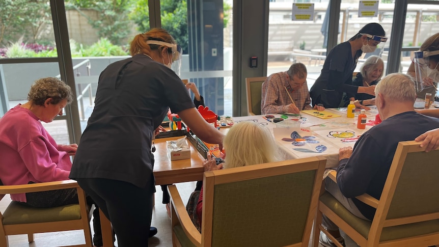 Staff in blue uniforms assist several elderly people sitting around a large table, painting pictures.