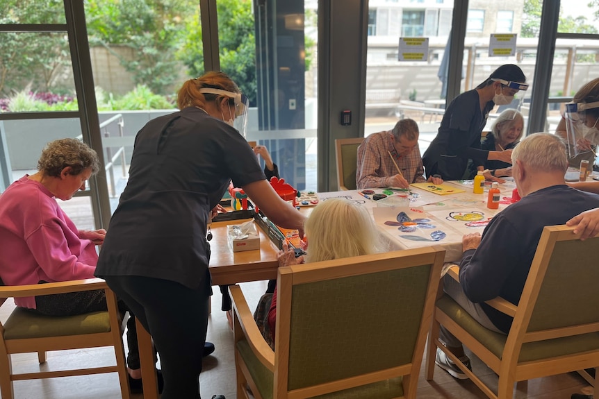 Staff in blue uniforms assist several elderly people sitting around a large table, painting pictures.