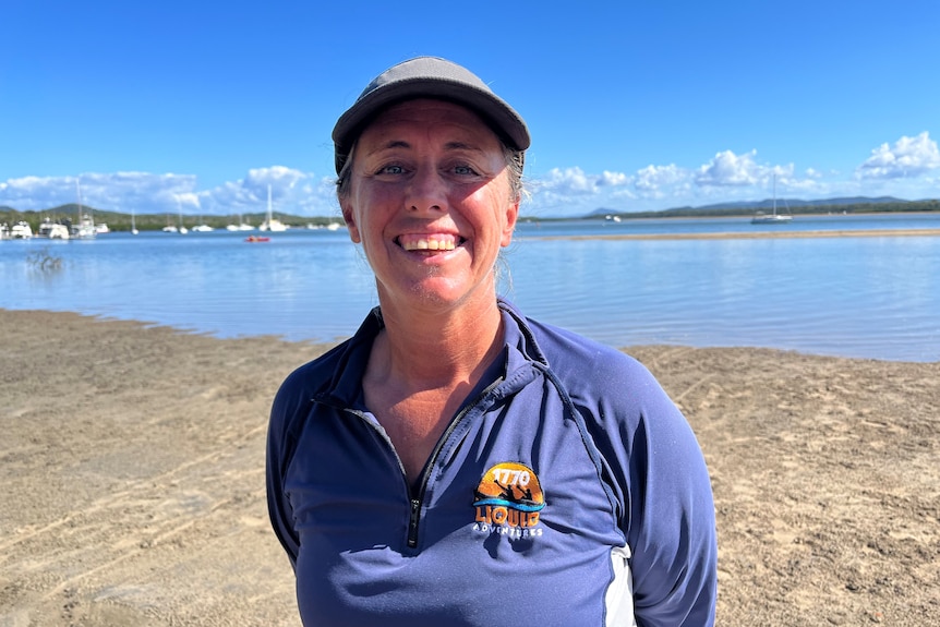 A woman in a cap and blue t-shirt stands smiling on the beach