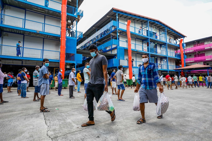 Men in face masks with bags of groceries walk through a courtyard surrounded by brightly coloured buildings