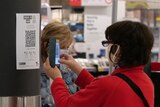 A woman in a red jacket and a face mask scans a QR code at a supermarket.