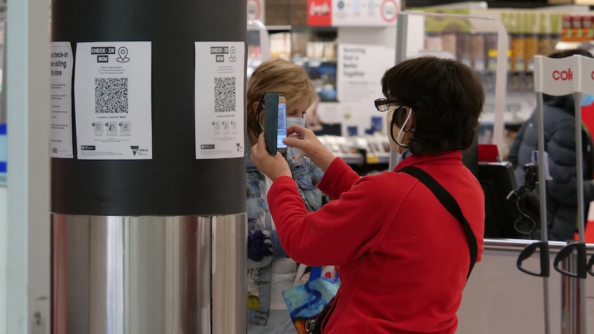 A woman in a red jacket and a face mask scans a QR code at a supermarket.