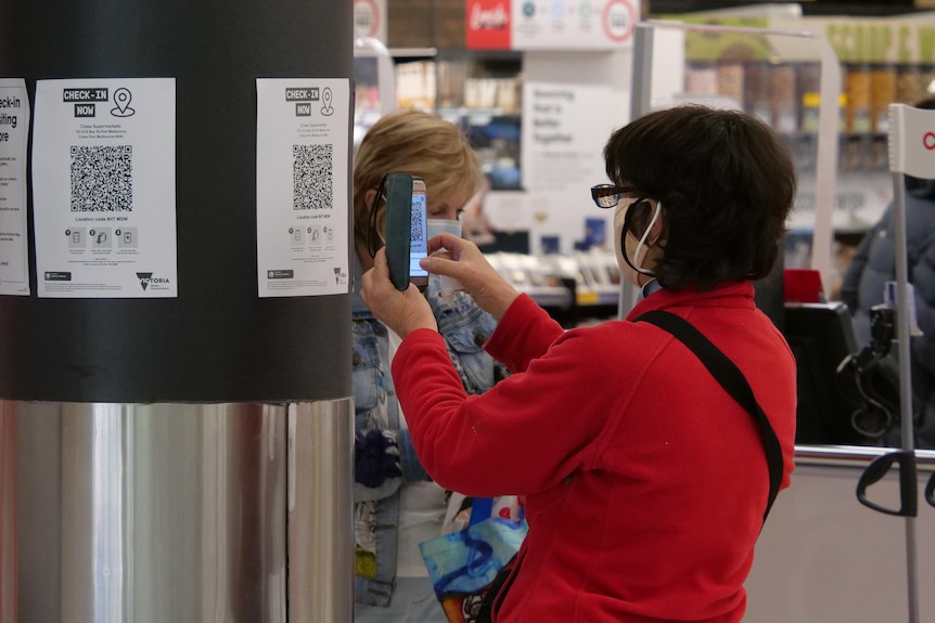 A woman in a red jacket and a face mask scans a QR code at a supermarket.