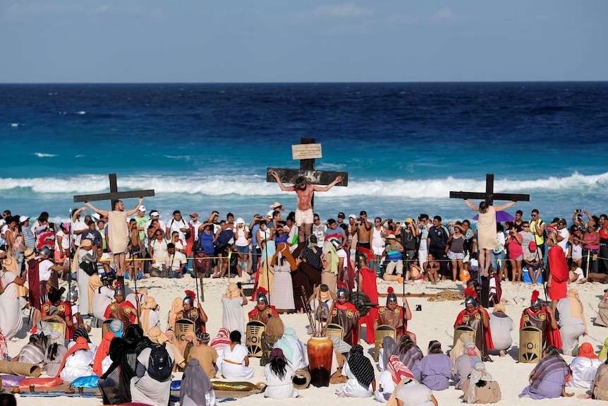 Three men are nailed to the cross as onlookers take photographs on the beach in Cancun