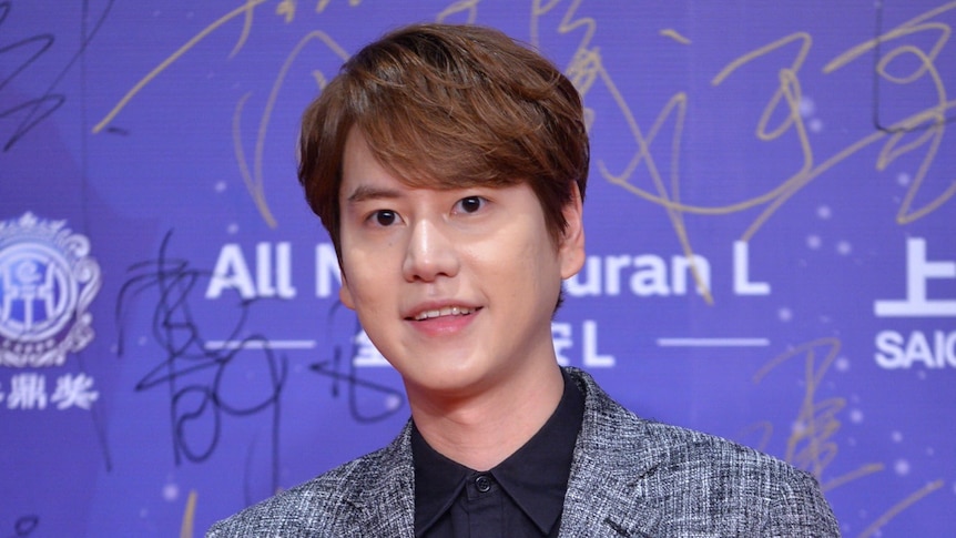 Kyuhyun in front of purple background posing with his hands held together.