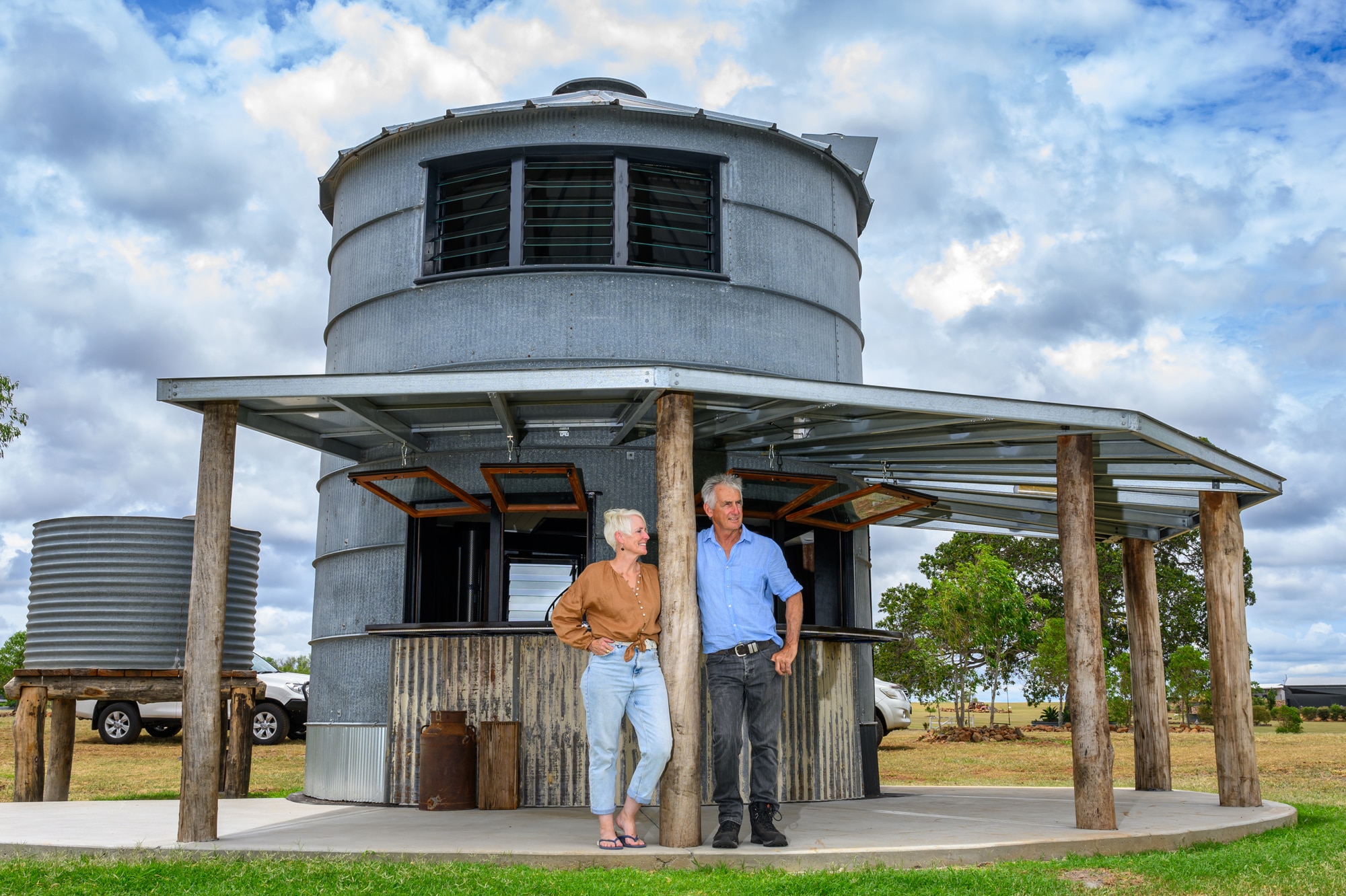 A woman with brown shirt and jeans and a man with blue shirt and jeans stand under a veranda attached to their silo hotel
