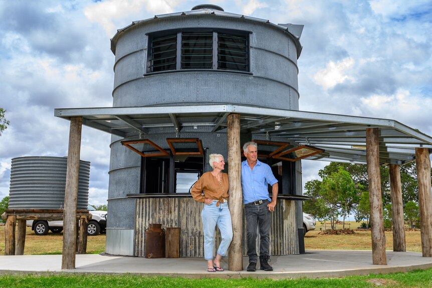 A woman with brown shirt and jeans and a man with blue shirt and jeans stand under a veranda attached to their silo hotel