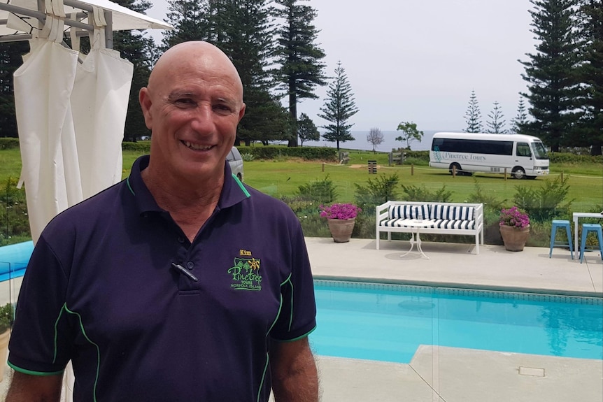 Un hombre de mediana edad sonriente está parado frente a una piscina con árboles y un minibús al fondo.