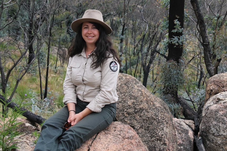 A woman in rangers uniform sits on a boulder in bushland.