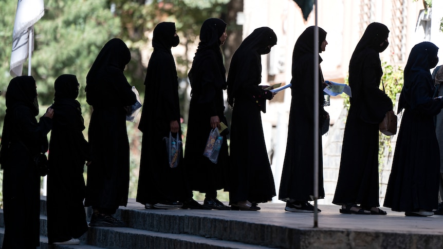 Afghan female students stand in a queue dressed in black abiyas and hijabs.