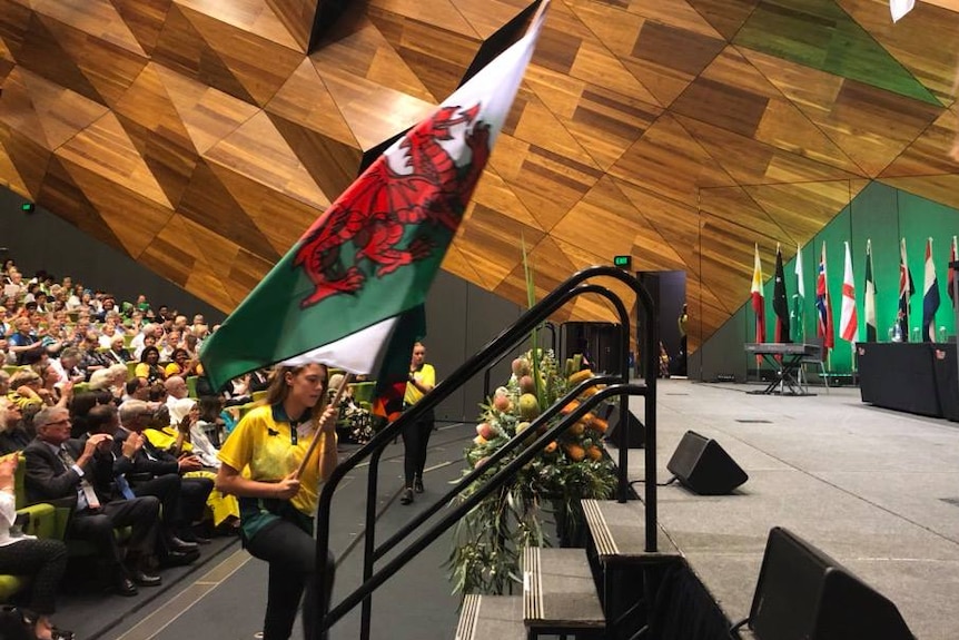 A young female flag bearer carrying a Welsh flag climbs the steps to a stage at a busy auditorium.