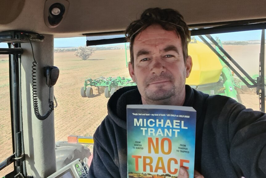 A selfie of a middle-aged man inside a tractor, holds book, wears grey hoodie, sunglasses on head, slight smile, unshaven face.