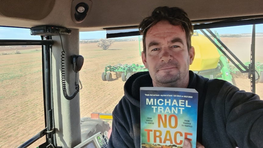 A selfie of a middle-aged man inside a tractor, holds book, wears grey hoodie, sunglasses on head, slight smile, unshaven face.