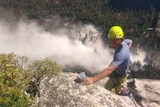 A man wearing a yellow helmet is climbing down a cliff with smoke and a forest below him.