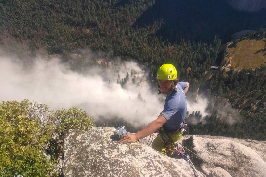 A man wearing a yellow helmet is climbing down a cliff with smoke and a forest below him.