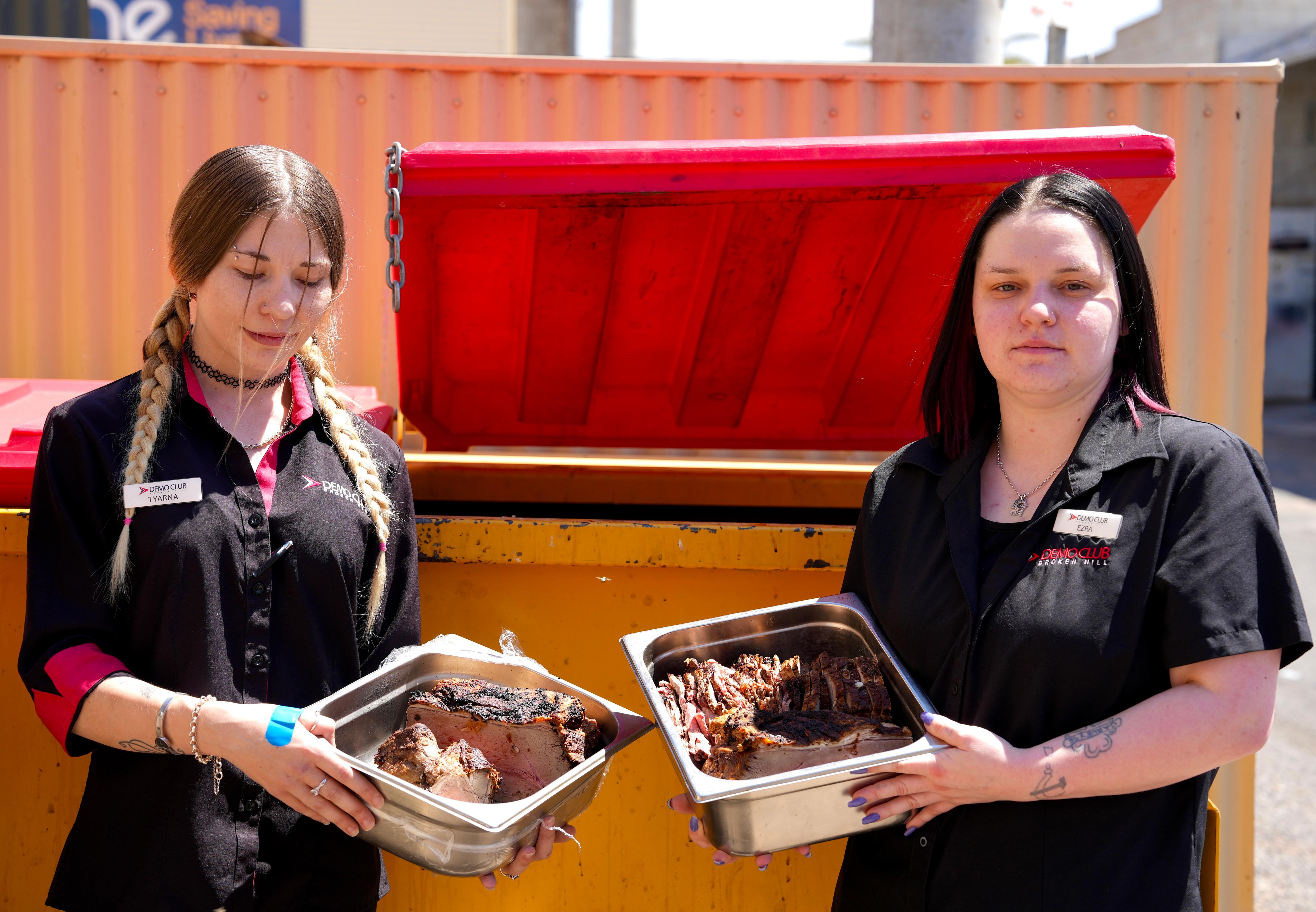 Dos mujeres se paran frente a un contenedor de basura sosteniendo bandejas de carne, ambas usan camisas negras con cuello rojo.