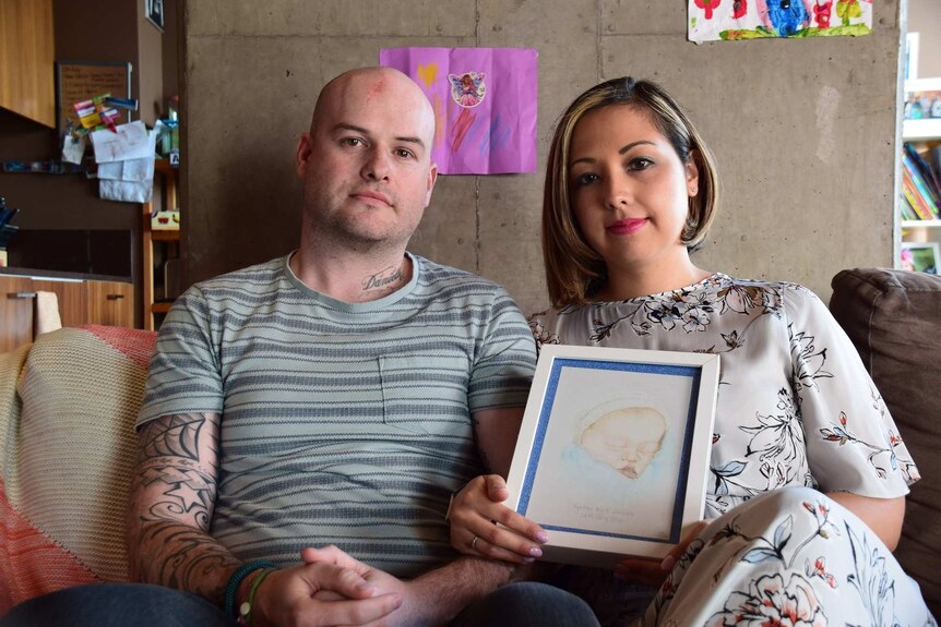 A man and woman sit next to each other, looking straight ahead. The woman holds a portrait of their stillborn son.