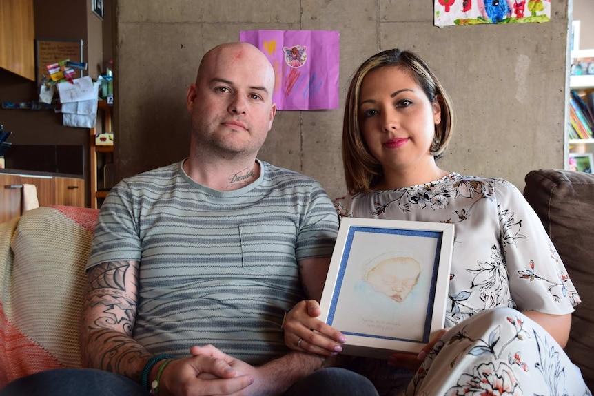 A man and woman sit next to each other, looking straight ahead. The woman holds a portrait of their stillborn son.