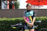 A woman wearing a Cycling Mums Australia jersey leans against her bike in front of a hedge.
