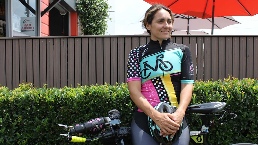 A woman wearing a Cycling Mums Australia jersey leans against her bike in front of a hedge.