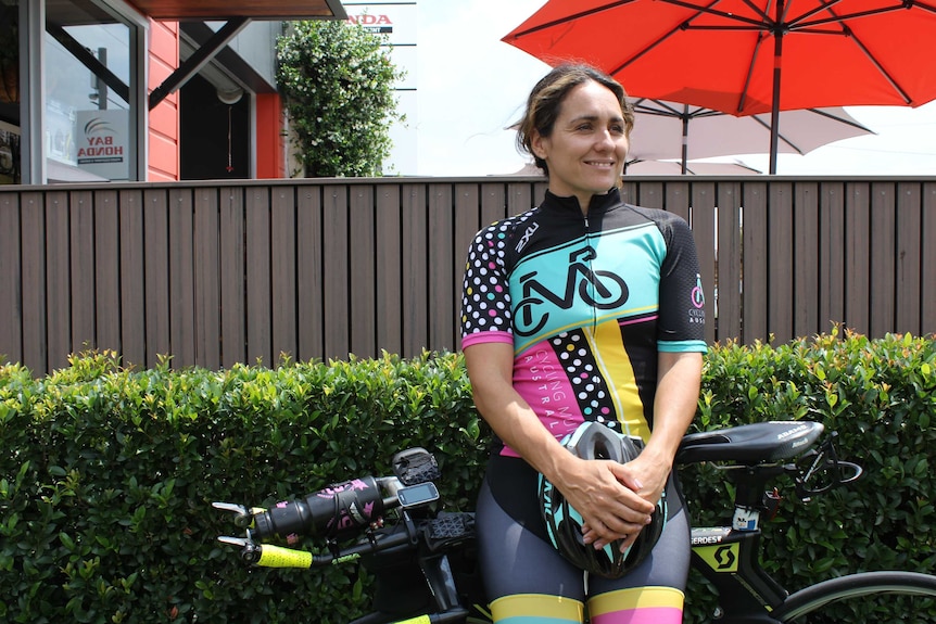 A woman wearing a Cycling Mums Australia jersey leans against her bike in front of a hedge.
