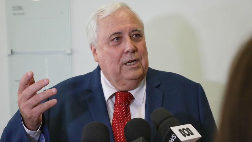 Clive Palmer, wearing a red tie, gestures while speaking into microphones in a hallway.