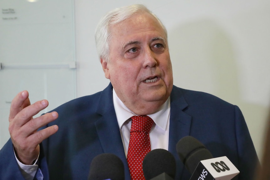 Clive Palmer, wearing a red tie, gestures while speaking into microphones in a hallway.