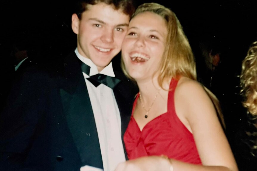 Kym, right, wears a red dress and smiles with Troy, left, who wears a tux and beams at the camera in this nighttime image.