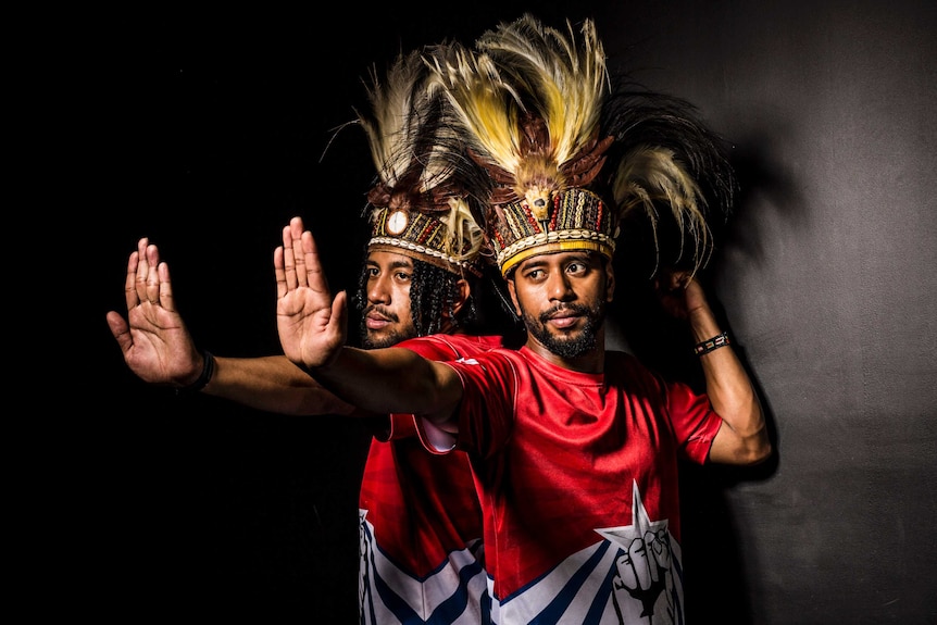 Mid-shot colour photograph of West Papua dancers and brothers Yoshua and Sam Roem against a black wall with a spotlight.