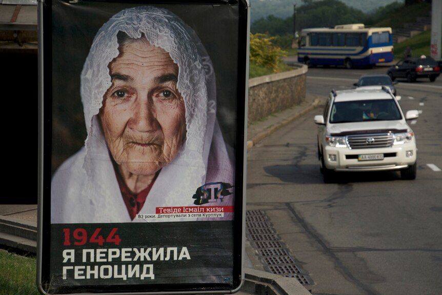 A billboard featuring an old woman displays the 1944 "I survived the genocide" poster.