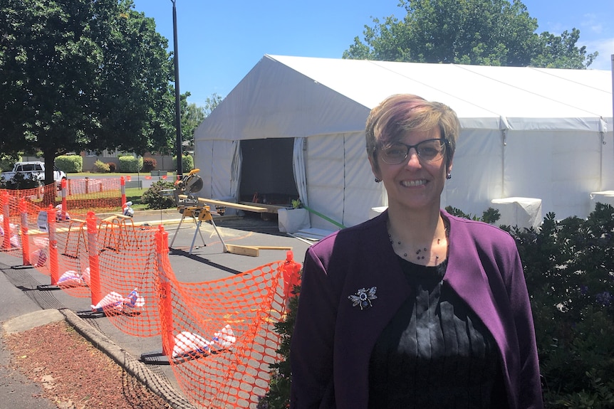 A woman with short blonde hair in a purple jacket smiles at the camera with a white tent behind her