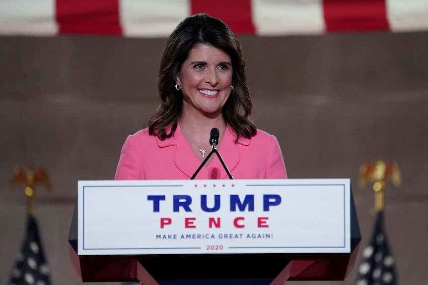 A woman in a pink suit smiles behind a lectern that says "Trump-Pence: Make America Great Again" 