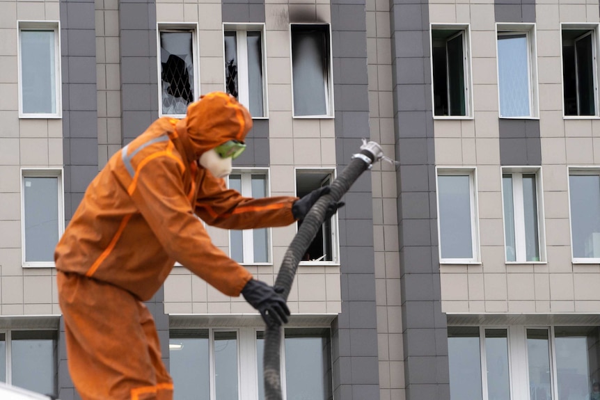 A person in an orange hazmat suit and black gloves handles a firehose, outside a building with several blackened windows