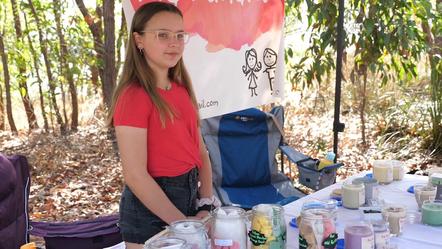 girl with glasses and red top stands selling candles in an area surrounded by trees.