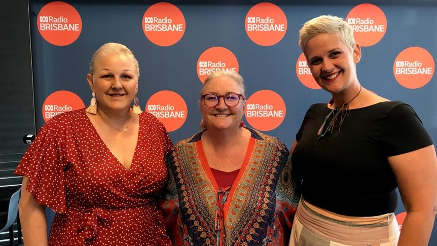 Shane Richards (L), Kelly Higgins-Devine and Tanya Allan stand smiling in front of the ABC Radio Brisbane wall.