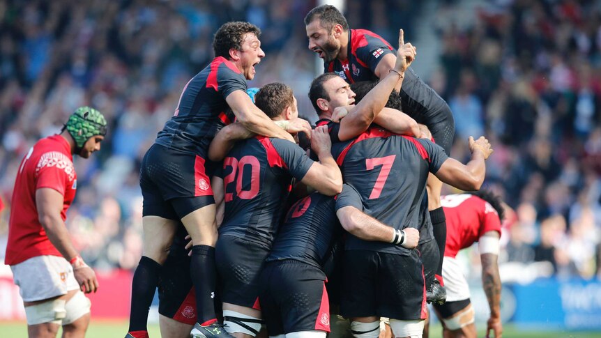 A group of male rugby union players join in a large circle as they embrace to celebrate winning a match.